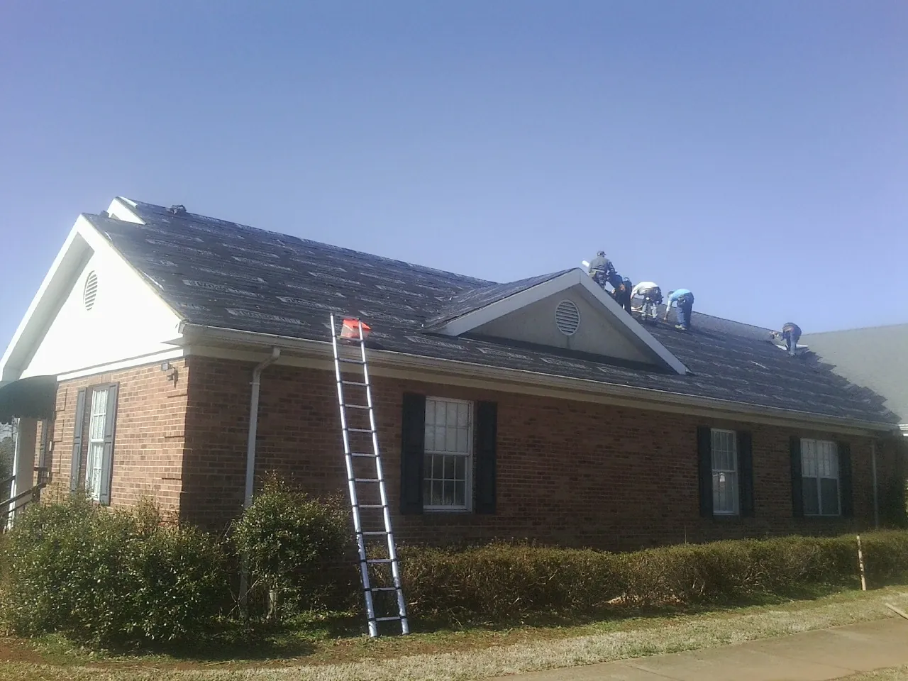 Crew tearing shingles off a brick ranch roof with a ladder set — Emergency Roof Repair work by our crew in Nanty Glo