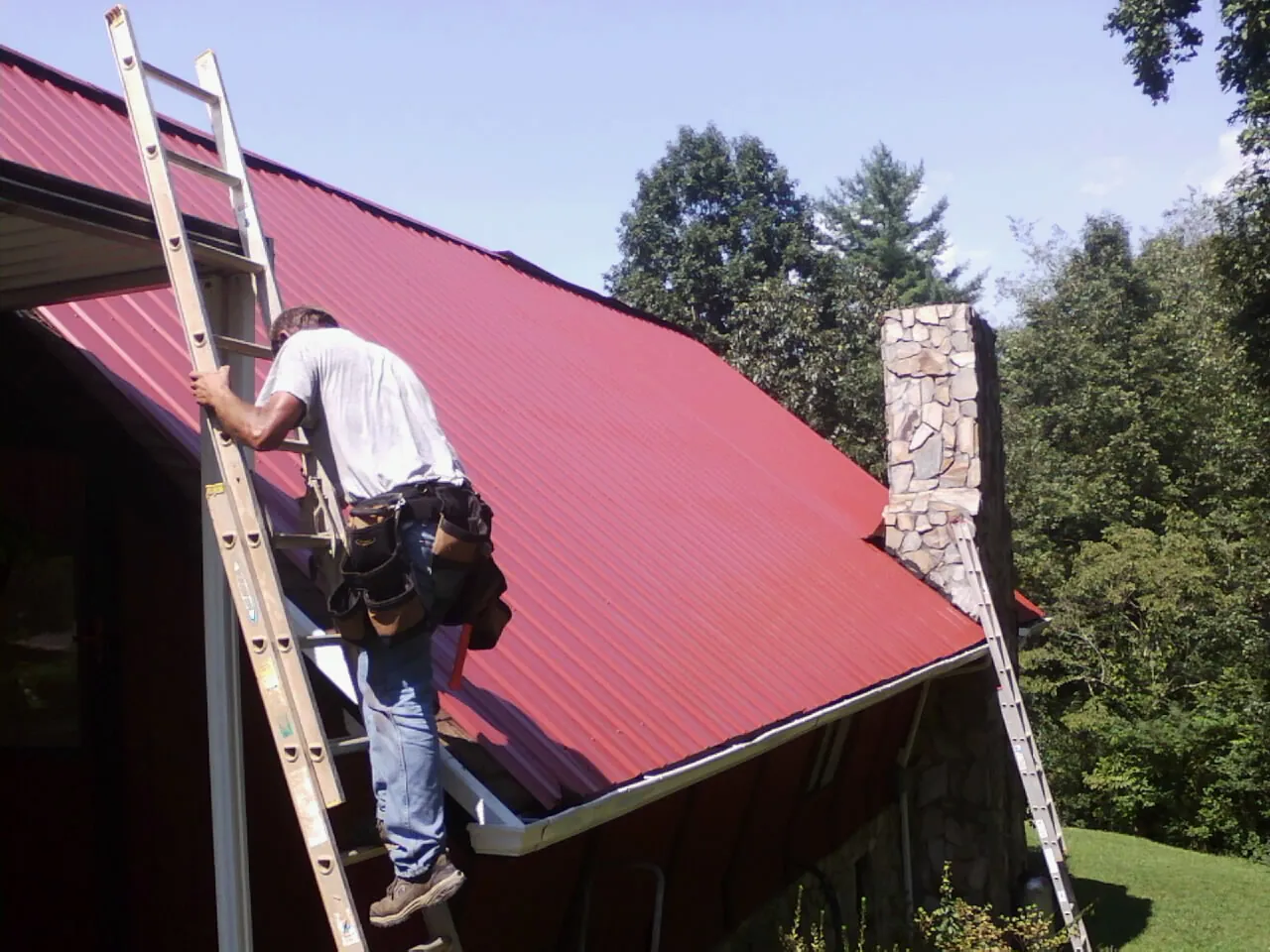 Roofer on a ladder beside a red metal roof and stone chimney — Soffit & Fascia Repair by Roofing Brothers in Nanty Glo